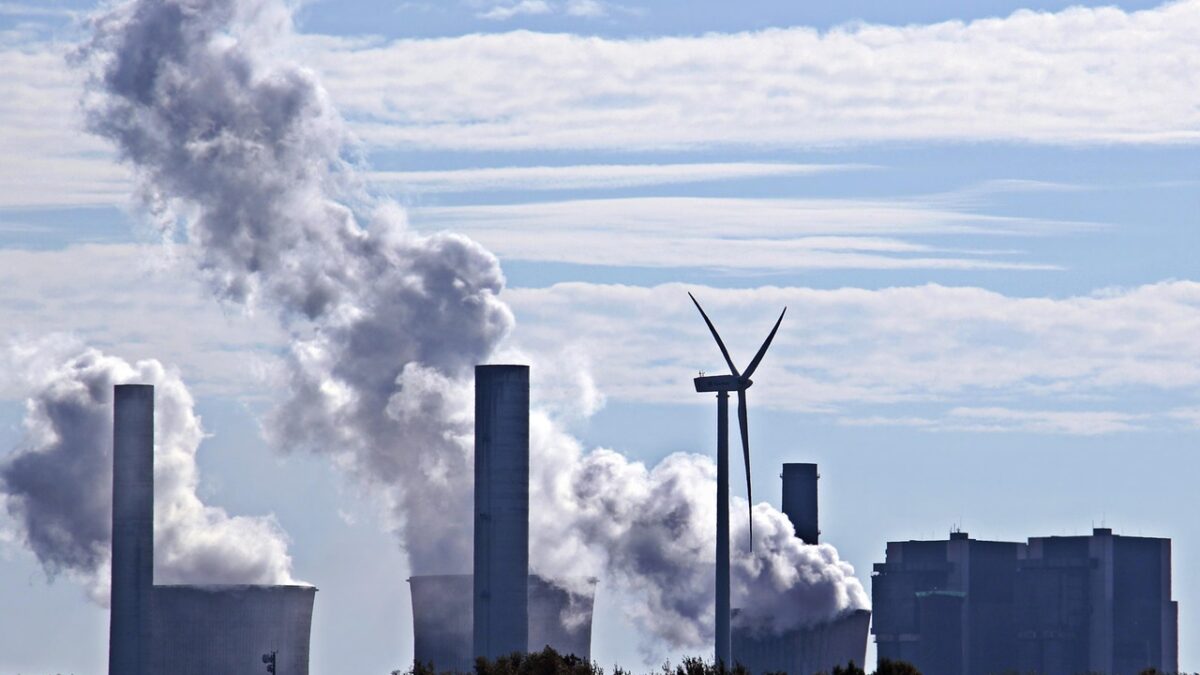 Centrale électrique aux nombreuses cheminées et tours de refroidissement fumantes, avec une éolienne au centre, des arbres au premier plan et un ciel bleu partiellement nuageux.