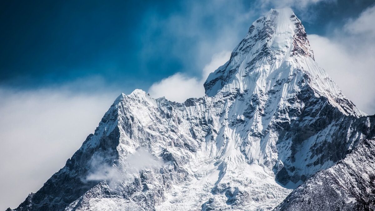 Haute montagne enneigée au sommet pointu, avec des crêtes rocheuses et des coulées de glace sous un ciel bleu nuageux.